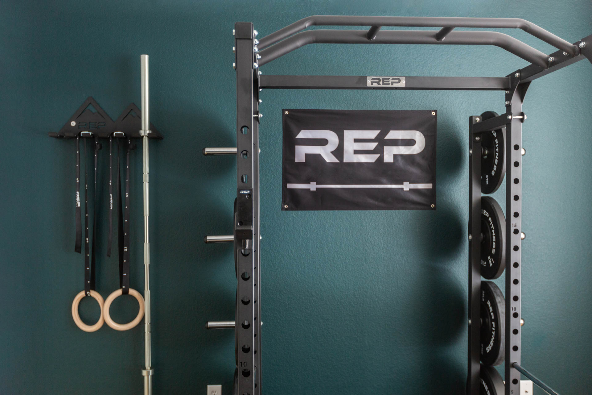 Home gym featuring a PR-1100 rack and a REP Multi-Use Wall Storage unit storing a barbell and gymnastic rings.